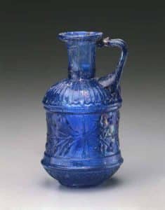 Cylindrical jug dating from the first half of the first century from Syria or Palestine, it is translucent royal blue with a brownish-green streak at the rim, created with medium thin glass and sporting one very large elongated bubble in the neck. Rim folded outward, upward, and inward. The cylindrical neck tapered slightly downward. Convex shoulder and convex bottom joined by a cylindrical body. Bifurcated handle applied to shoulder and attached to the rim with projecting thumb rest above. Gift of Edward Drummond Libbey.