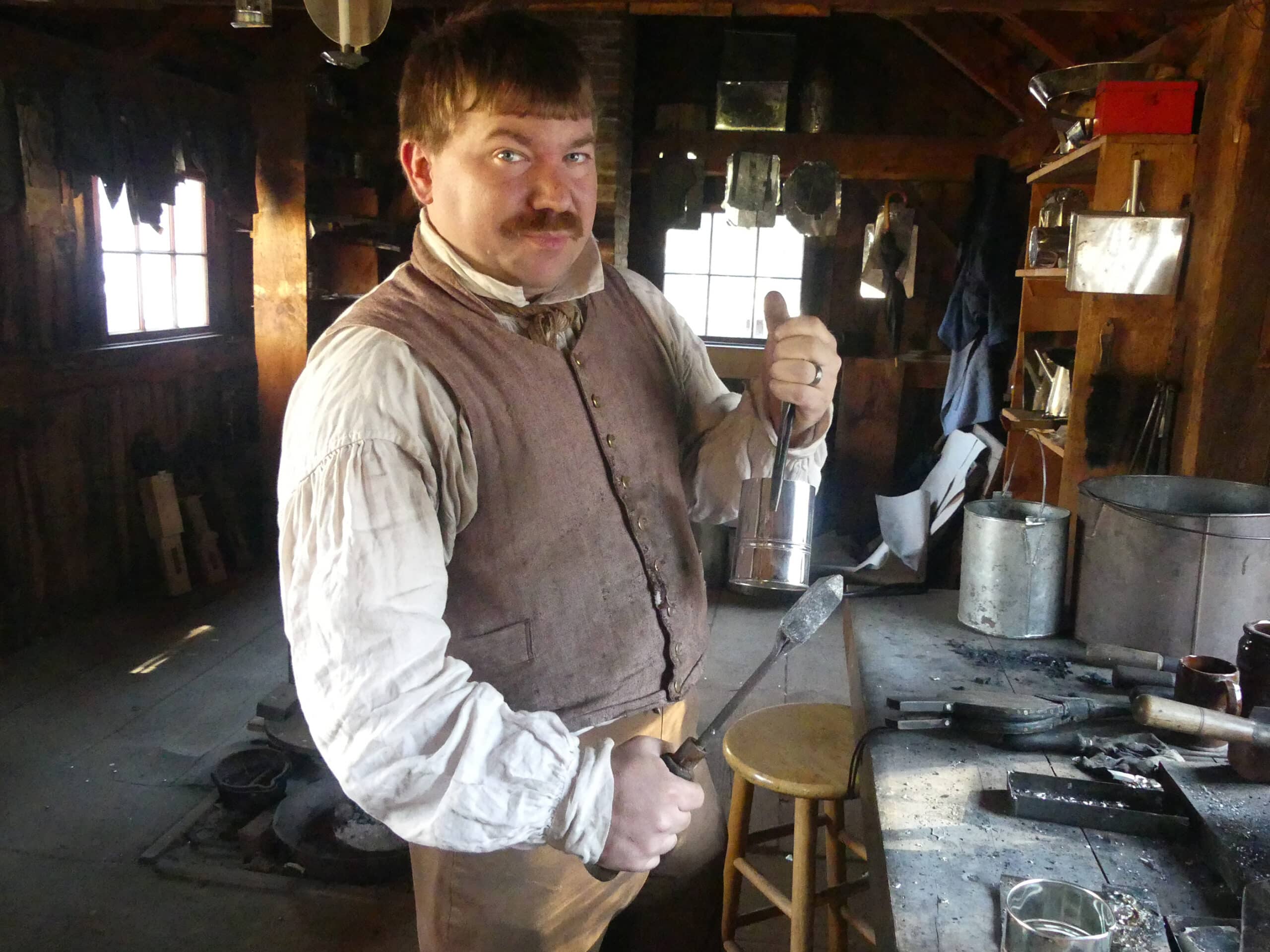 Here, the body of the cup was shaped and has been soldered to keep its shape and to make it watertight. The tool in his right hand is the soldering copper which has been heated in the charcoal fire to melt the solder.