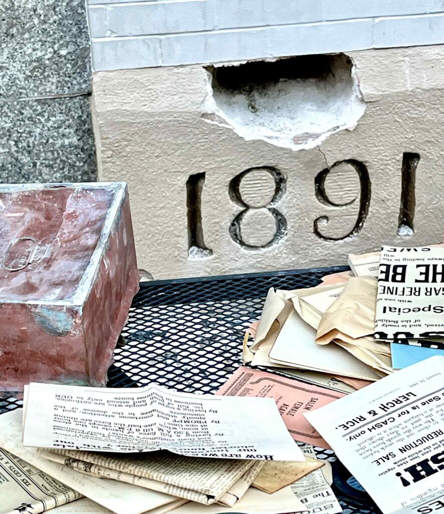 A grouping of the time capsule box and its contents from 1891 around the cornerstone of the “New Bee Hive."