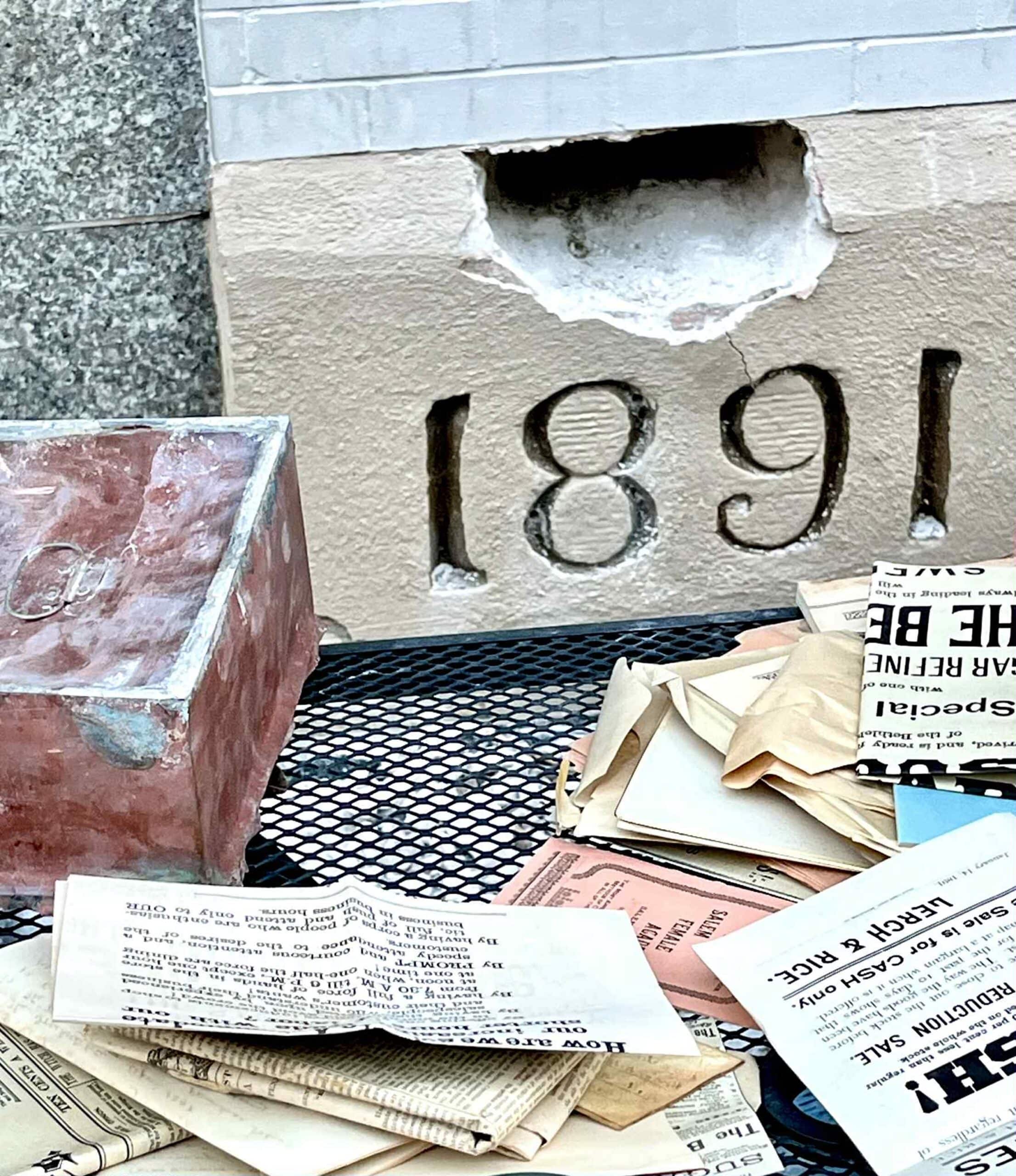 A grouping of the time capsule box and its contents from 1891 around the cornerstone of the “New Bee Hive."